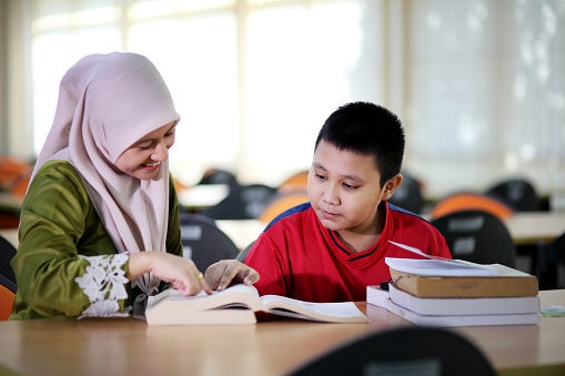 A Muslim female adult is guiding a young boy on his studies in library.