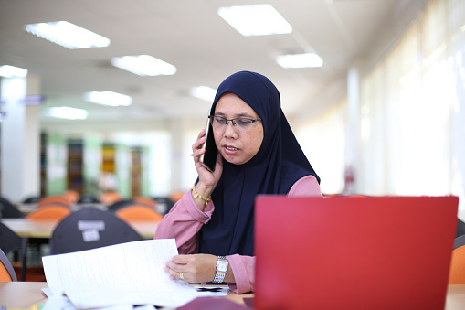 Muslim female adult spending time talking on the phone while doing research in the library.