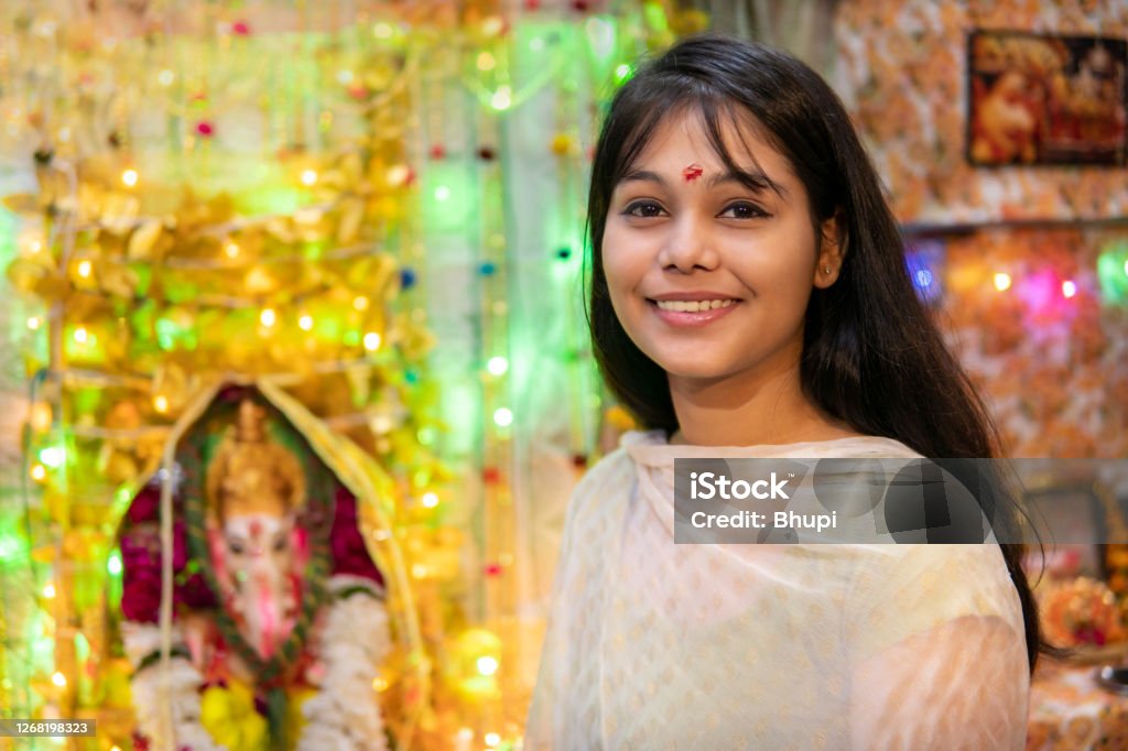 happy Indian girl celebrating ganesh chaturthi festival. Indoor image of beautiful, happy Indian girl sitting near ganesha statue and celebrating ganesh chaturthi  festival at home. she is wearing traditional Indian dress and looking at camera with toothy smile Ganesh Chaturthi Stock Photo