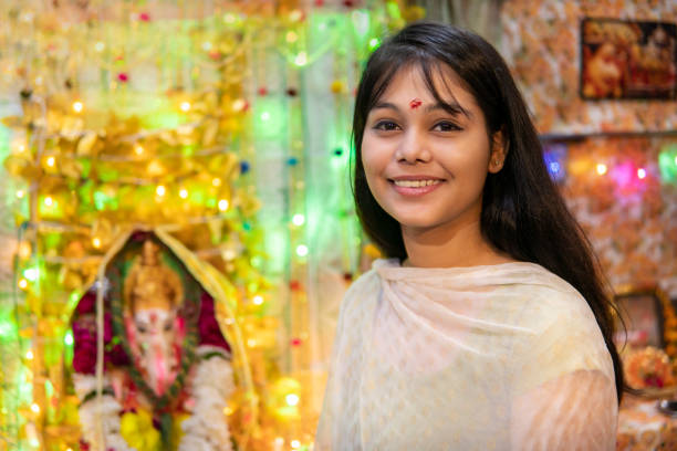 happy Indian girl celebrating ganesh chaturthi festival. Indoor image of beautiful, happy Indian girl sitting near ganesha statue and celebrating ganesh chaturthi  festival at home. she is wearing traditional Indian dress and looking at camera with toothy smile beautiful traditional indian girl stock pictures, royalty-free photos & images
