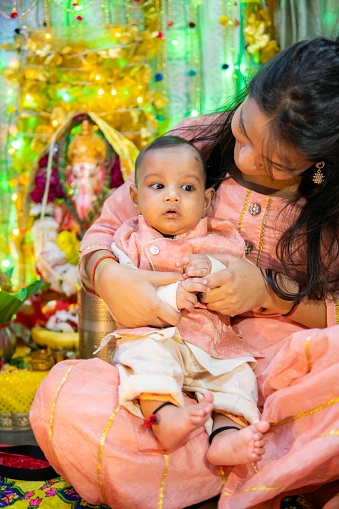 Indoor image of beautiful, happy Indian young woman sitting near ganesha statue with her cue little nephew and celebrating ganesh chaturthi festival at home. She is wearing traditional Indian clothes.