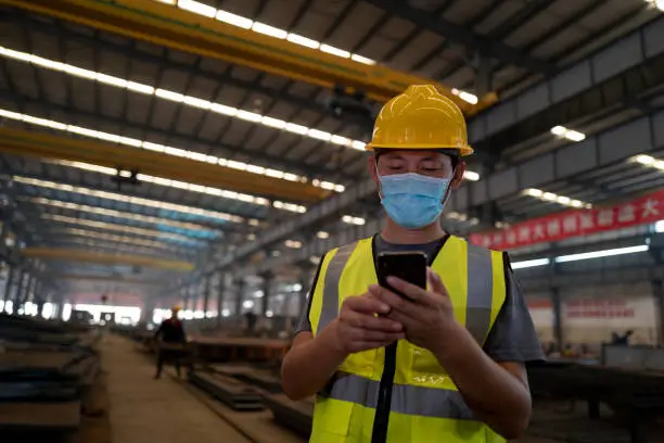 Photo of Asian male engineer wearing face mask using mobile phone at construction site