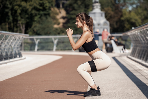 Concept of healthy and active lifestyle. Profile side view of serious and young adult woman making sport training outdoors alone, squat with resistance band equipment, holding hands in front