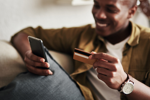 Cropped shot of a handsome young man smiling while using a smartphone and a credit card to shop online in his living room at home