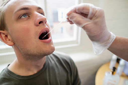 Extreme close-up of a mature caucasian health professional wearing a mask and visor using a cotton swab in a young male's mouth.