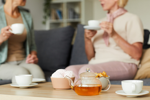 Close-up of teapot with tea with cups on the table and two women talking to each other on the sofa in the background