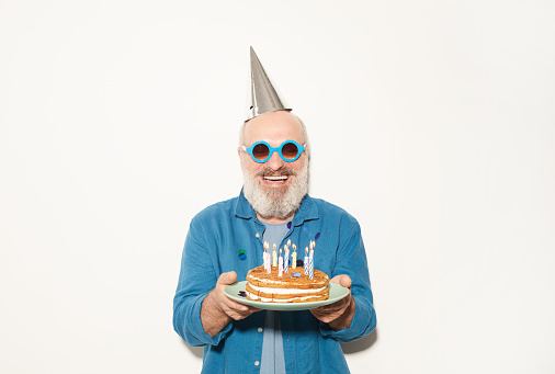 Portrait of senior man in party hat and sunglasses holding birthday cake and smiling at camera against the white background