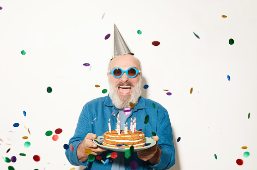 Portrait of excited senior man in sunglasses smiling at camera while standing under the confetti and holding cake isolated on white background