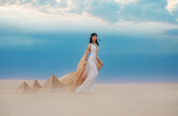 istock young beautiful woman model image Queen Cleopatra walks in desert Egyptian pyramids. Creative retro traditional gold jewelry bob hair cut. White satin long vintage dress, orange silk cape fly train 1268232986