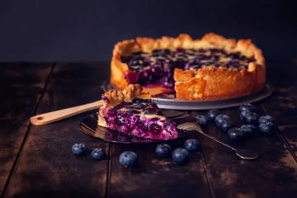 Rustic shortbread pie with blueberries in sour cream filling on a wooden background with a piece cut off on a saucer and a spoon