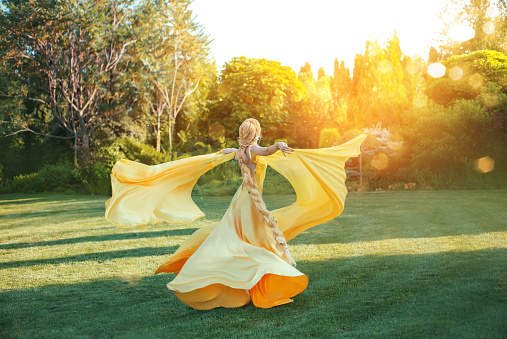 Fantasy happy woman queen dancing. hand raised. yellow dress long train. Silk cloak fluttering flying in wind. hairstyle long blonde braid hair. Art photo image of Princess back, rear view. divine sun
