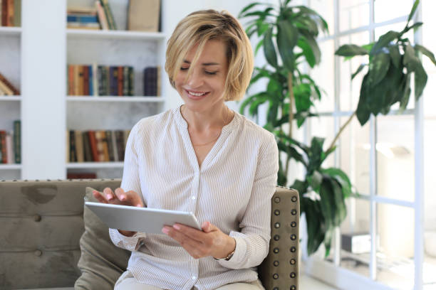 istock Cheerful middle aged woman sitting on sofa, using computer tablet apps, looking at screen,reading good news in social network, shopping or chatting online. 1268242161