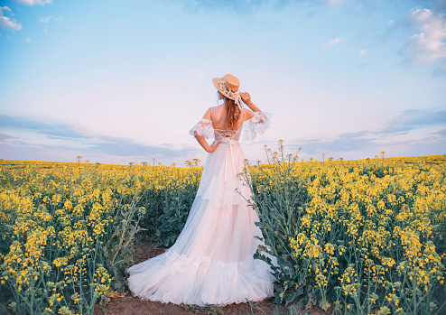 Art Village authentic beauty retro lady, enjoy nature blooming yellow rape field. Concept harmony. Long white pink vintage boho wedding dress train. Straw hat boater flowers. woman turned away back