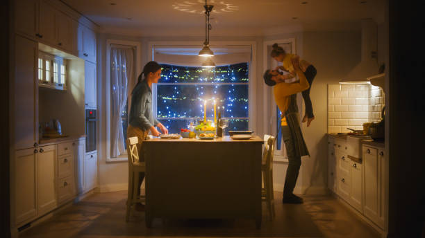 happy family of three cooking and having dinner together. mother prepares food, father hugs cute little girl. festive table in cozy kitchen - child party group of people little girls imagens e fotografias de stock