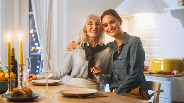 キッチンで美しい女性の多世代。高齢の母親と成熟したドーガーは、テーブルに座って抱擁し、カメラで楽しんで微笑みます。世代の再会と素敵な家族の夕食。 - grandmother senior adult family domestic kitchen ストックフォトと画像