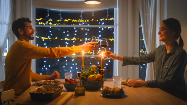 Happy Young Couple In Love Have Romantic Dinner Toasting Each Other With Glasses Of Wine Eating Tasty Meal In The Kitchen Celebrating Talking Beautiful Lovely And Wife Have Romantic Time Stock