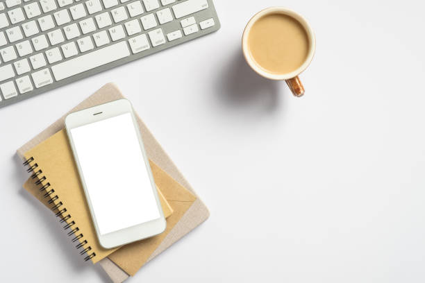 istock Modern office desk table with pc keyboard, coffee cup, paper notebook, smartphone mockup with blank screen on white background. Flat lay, top view. Minimal style workspace. 1268245213