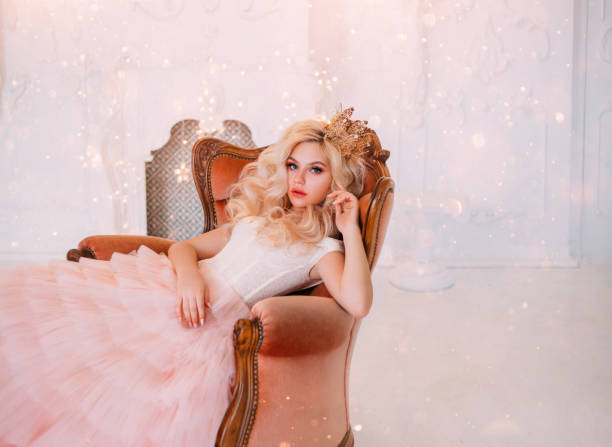 Young beautiful queen woman resting in medieval armchair. beauty face. New Year garland scenery glare sparks bright. Backdrop white classic room interior. Hairstyle princess girl blonde long wavy hair Young beautiful queen woman resting in medieval armchair. beauty face. New Year garland scenery glare sparks bright. Backdrop white classic room interior. Hairstyle princess girl blonde long wavy hair prom queen stock pictures, royalty-free photos & images