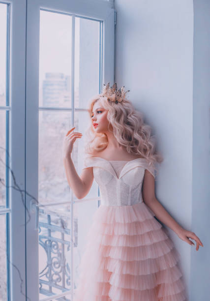 beautiful young sad fairy tale cute queen woman stand near vintage antique window. girl princess blond long wavy hair. luxury royal tender pink tulle dress. medieval gold crown. white room interior - cute indoors long hair real people imagens e fotografias de stock