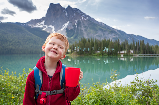 Happy Young Cute Redhead Boy smiling  at Emerald Lake in Summer. Yoho National Park, Canada. He is wearing a bear bell, an hydration backpack and he is holding a red coffee cup.