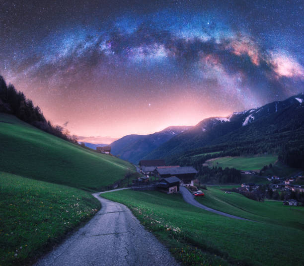 Photo of Arched Milky Way over the rural mountain road in summer in Italy. Beautiful night landscape with starry sky, milky way arch, winding road in mountain village, hills, green meadows and buildings. Space
