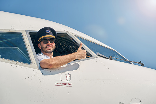 Joyous bearded Caucasian young airline pilot using a thumbs-up gesture in front of the camera