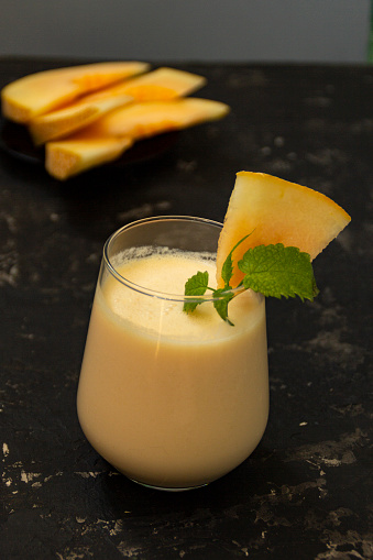 Melon milk shake on a black background. Ripe sliced melon is lying nearby. The smoothie glass is decorated with a sprig of mint and melon crumbs. Summer healthy food concept.
