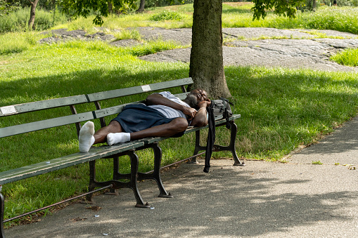 New York, NY, USA - August 22, 2020: A man in socks sleeps on a bench in the shade near the north ballfields on the upper east side of Manhattan's Central Park.
