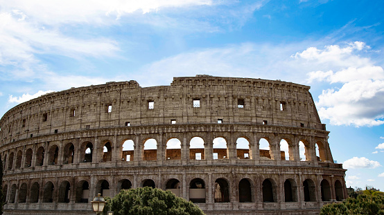 High angle view of Coliseum in downtown Rome at dusk, surrounded by ancient buildings located in the nearby Roman Forum, along with old churches, domes, bell towers and rooftops. Plenty of people walking in a street just in front of the monument.