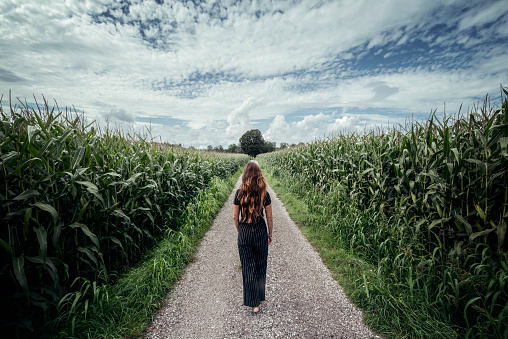 Austria. A woman with red hair walking on a long straight path. Dramatic sky and agriculture field on both sides.