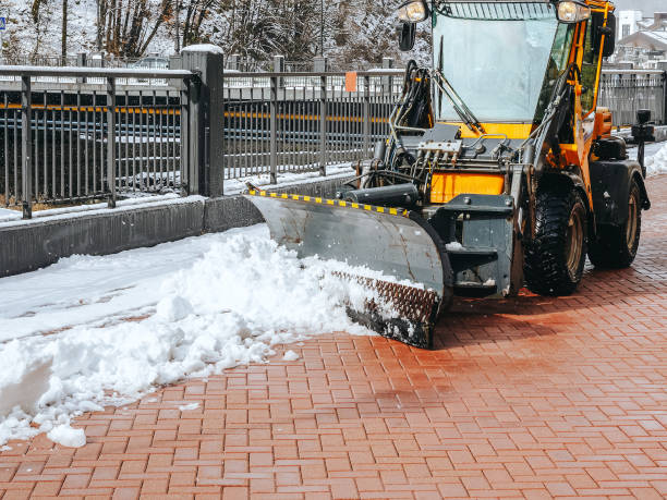 traktor räumt schnee von pflasterplatten entlang eines schwarzen metallzauns auf der straße - winterdienst stock-fotos und bilder