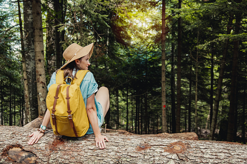 Woman with backpack and hat sitting on the fallen tree and relaxing