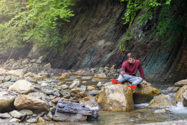istock A man looks at the phone while sitting on a stone on a mountain river. 1268327418