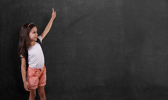 Little cute girl is pointing with her hand in front of chalkboard.