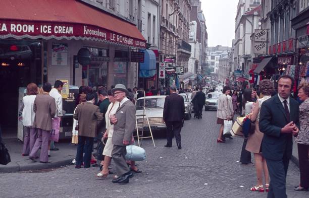 Paris, Il de France, France, 1975. Street scene in Paris. Parisians and tourists stroll and shop through the streets of an old district of the main French town. Also: shops, advertising, road traffic, cars and buildings.