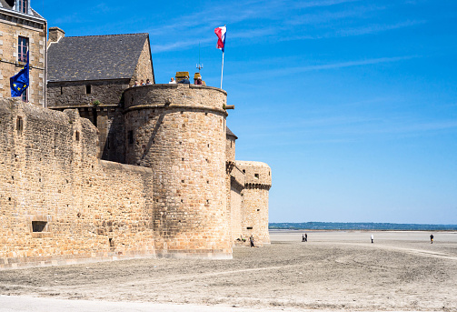 Beauvoir, France.  May 12, 2019.  Section of the castle of Mont St. Michel with flags blowing and people walking on the beach.  Sunny day with blue skies.