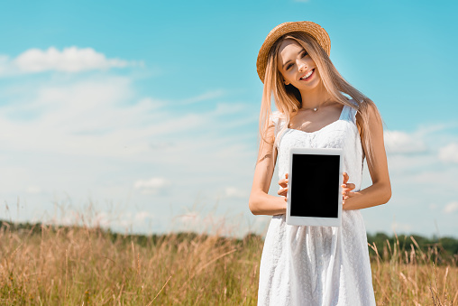 blonde woman in straw hat and white dress showing digital tablet with blank screen while standing in filed