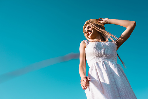 low angle view of blonde woman in white dress touching straw hat against blue sky