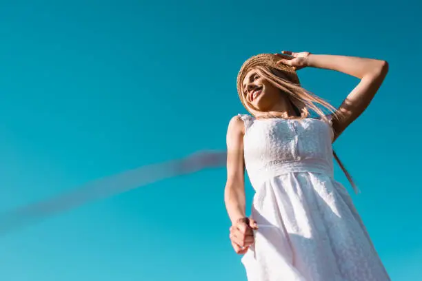 Photo of low angle view of blonde woman in white dress touching straw hat against blue sky
