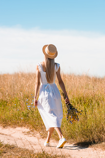 back view of blonde woman in white dress and straw hat walking on road in field with bouquet of wildflowers