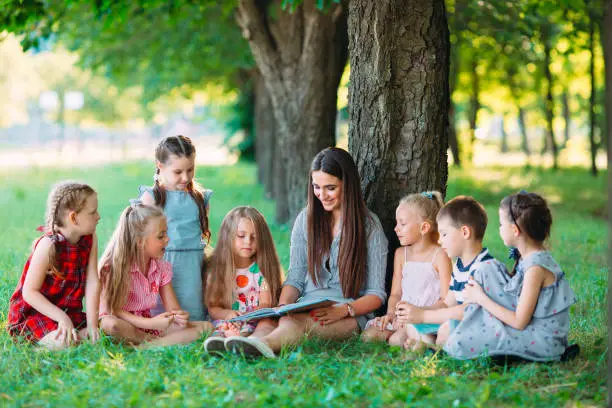 Photo of Children and education, young woman at work as educator reading book to boys and girls in park.