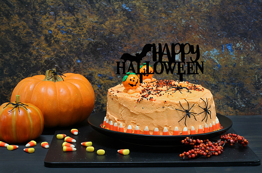 Happy Halloween cake with orange frosting, Happy Halloween banner, pumpkins, candy corn and spiders in a low light setting.