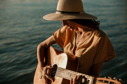 Portrait of pensive attractive Caucasian woman in a floral dress and with hat sitting on the shore next to the river and playing the guitar. Peaceful and in a harmonious music vibe.