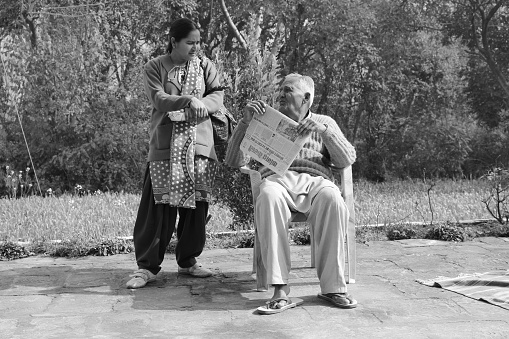 Kalarkalan, Kangra, Himachal Pradesh, India - February 11,2015: A woman having a discussion with an old man in a village of Himachal Pradesh while he folds a newspaper he was reading while sitting on a chair with the backdrop of a farm. Kangra is largest populated district in Himachal Pradesh. Himachal Pradesh has 7 lakh persons aged 60 years and above, constituting 10.2% of its total population, which is higher than the national average of 8.6% (Census 2011) while elderly constitute 11.7 % of Kangra's population.