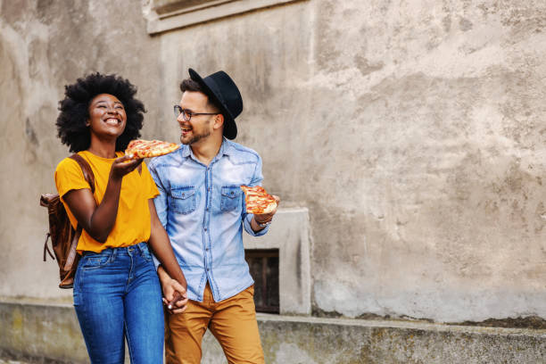 longitud completa de feliz pareja multirracial caminando al aire libre, cogidos de la mano y comiendo pizza. - couple black american culture african culture fotografías e imágenes de stock