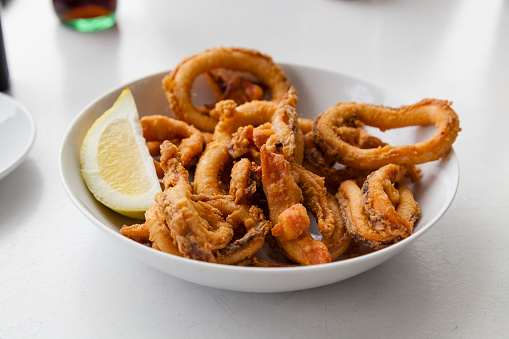 Food shot of a Spanish fried squid tapas dish with lemon and a glass of soda on a background bar table with copy space. Cantabria traditional Rabas snack.