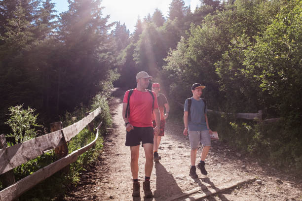 mature man and his sons, teen boys hiking, walking on a dirt road in the forest in summer - backlit, serious, having adventures, developing togetherness, bonding, being healthy, active, fit. - dirtroad imagens e fotografias de stock