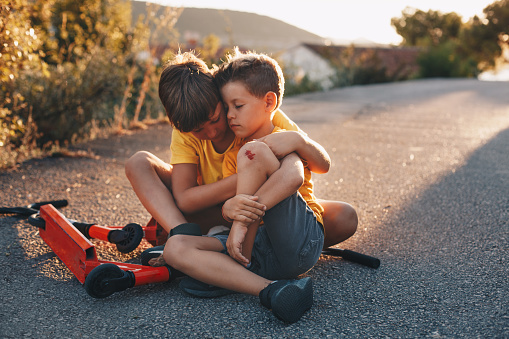 A younger brother was riding a scooter down the road and fell. He tore his knee and was bleeding. His big brother is there for him, hugging him and comforting him. Everything will be all right!