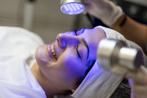 Skin Cosmetology. Woman beautician Doing Blue Light Therapy On face of pretty young woman