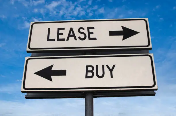 Photo of Lease vs buy. White two street signs with arrow on metal pole with word. Directional road. Crossroads Road Sign, Two Arrow. Blue sky background. Concept for own property versus borrow it.